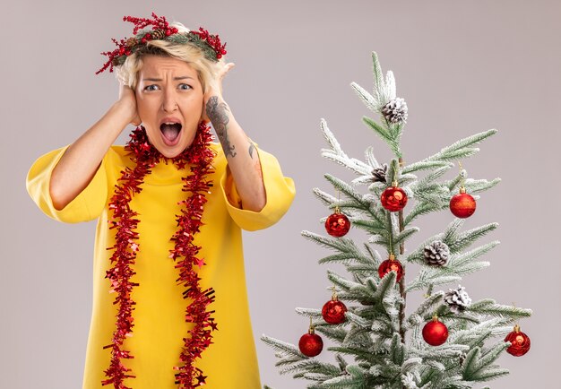 クリスマス ヘッド リースと見掛け倒しの花輪を身に着けている若いブロンドの女性が飾られたクリスマス ツリーの近くに立って、白い壁に孤立した頭に手を当てて叫び続けることを強調した