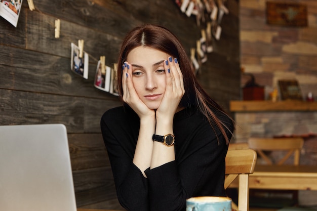Free photo stressed young caucasian female freelancer resting face on her hands looking at laptop screen in front of her with bored expression, feeling tired while working remotely at cafe. people and lifestyle