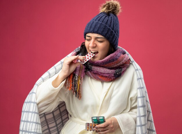 Stressed young caucasian ill girl wearing robe winter hat and scarf wrapped in plaid holding packs of capsules biting one of them with closed eyes isolated on crimson background