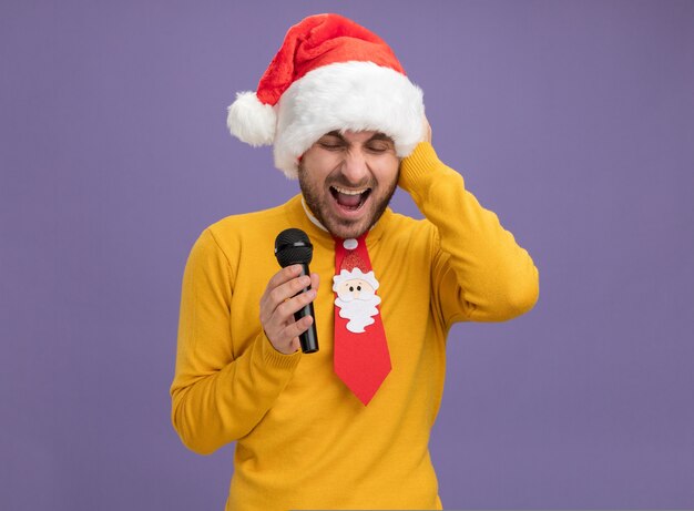 クリスマスの帽子とネクタイを身に着けている若い白人男性を強調し、コピースペースで紫色の背景に分離された目を閉じて叫んで頭に手を保ちます