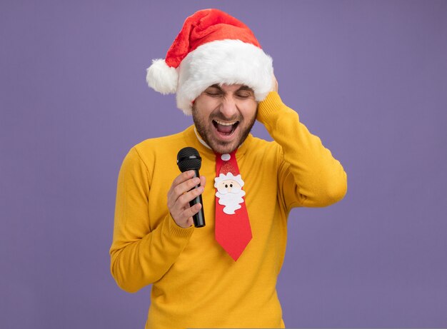 Stressed young caucasian man wearing christmas hat and tie holding microphone keeping hand on head screaming with closed eyes isolated on purple background with copy space