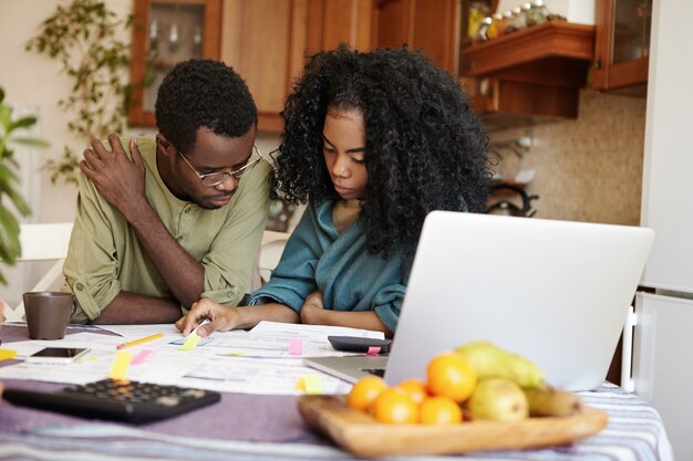 Free photo stressed young dark-skinned married couple looking frustrated while calculating domestic budget together, sitting at kitchen table with lots of papers and laptop computer, trying to save some money