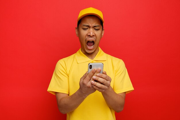 Stressed young delivery man wearing cap and uniform holding and looking at mobile phone screaming 