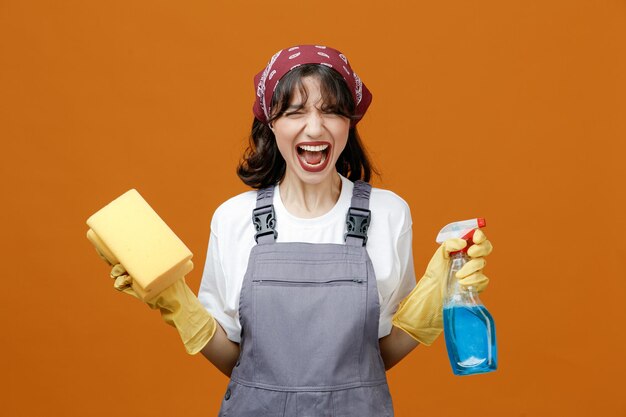 Stressed young female cleaner wearing uniform rubber gloves and bandana holding sponge and cleanser screaming with closed eyes isolated on orange background