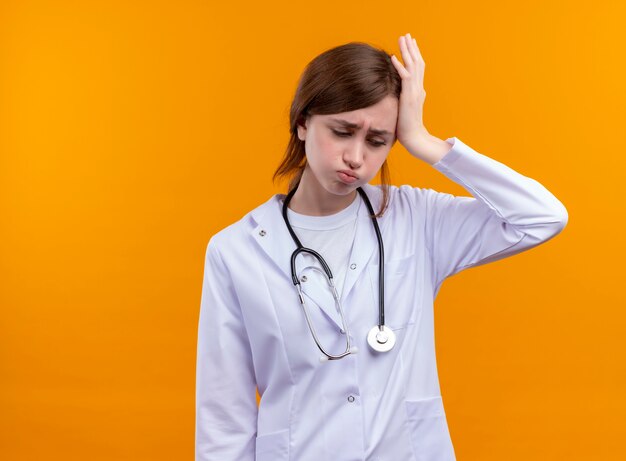 Stressed young female doctor wearing medical robe and stethoscope putting hand on head looking down on isolated orange wall with copy space