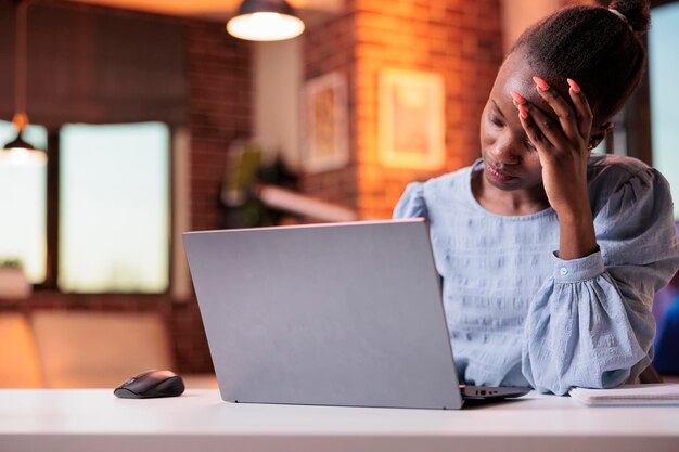 Stressed young female freelancer having headache and working on laptop, corporate employee burnout concept. Tired busy african american businesswoman, unhappy employee at workplace