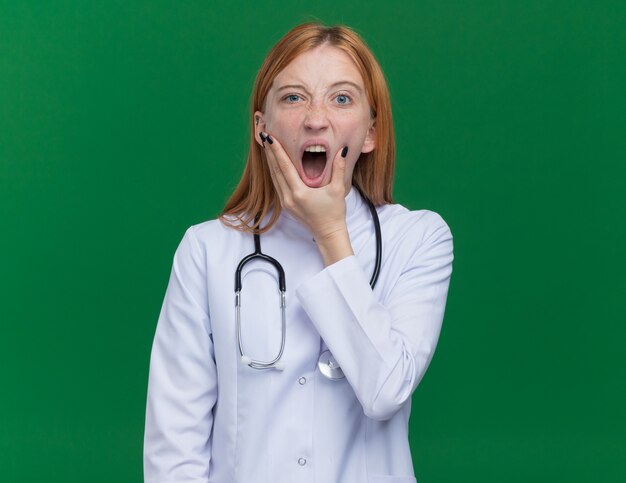 医療用ローブと聴診器を身に着けている若い女性の生姜医師がコピースペースで緑の背景に分離されたカメラを見て叫んで顎に手を置いている