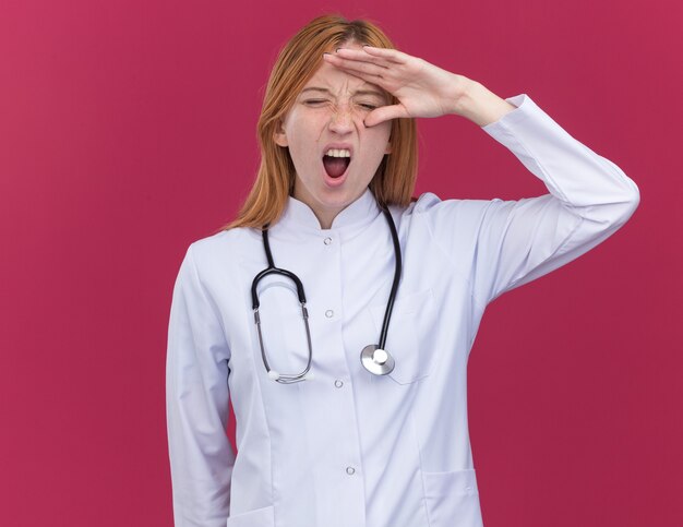 Stressed young female ginger doctor wearing medical robe and stethoscope keeping hand on forehead screaming with closed eyes isolated on crimson wall