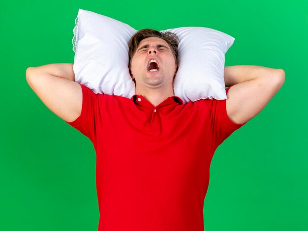 Stressed young handsome blonde ill man holding pillow under head screaming with closed eyes isolated on green wall