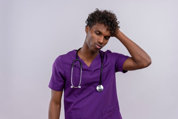 Подчеркнутый молодой красивый темнокожий доктор с кудрявыми волосами в фиолетовой форме со стетоскопом держит руку на голове с закрытыми глазами