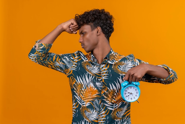 青い目覚まし時計を押しながらオレンジ色の背景に時間を示す葉プリントシャツの葉の巻き毛を持つ若いハンサムな浅黒い肌の男を強調