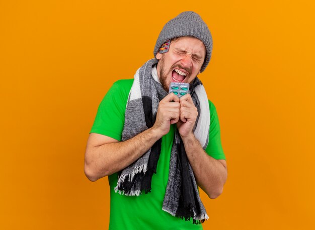 Stressed young handsome ill man wearing winter hat and scarf holding pack of capsules screaming with closed eyes with pack of capsules under hat isolated on orange wall