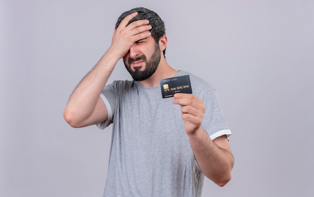 Free photo stressed young handsome man holding credit card and putting hand on head with closed eyes isolated on white wall