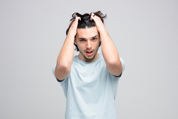 Stressed young handsome man keeping hands on head grabbing his hair while looking at camera isolated on white background