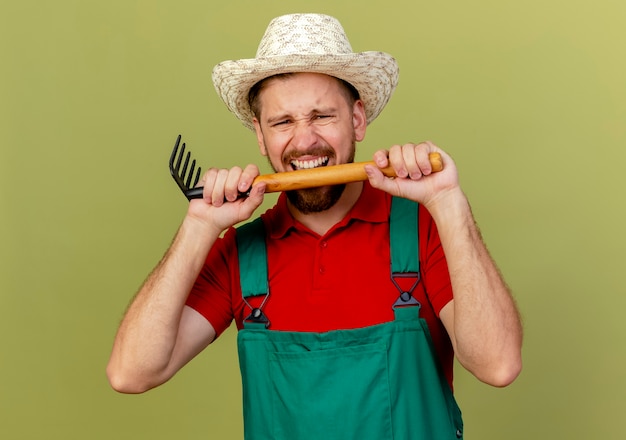 Stressed young handsome slavic gardener in uniform and hat holding and biting rake  isolated on olive green wall