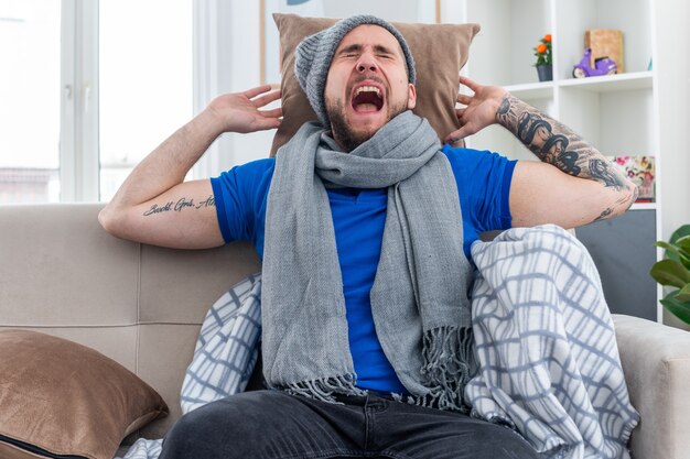 Stressed young ill man wearing scarf and winter hat sitting on sofa in living room holding pillow behind his head screaming with closed eyes