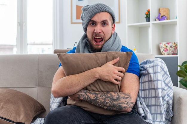 Stressed young ill man wearing scarf and winter hat sitting on sofa in living room hugging pillow  screaming