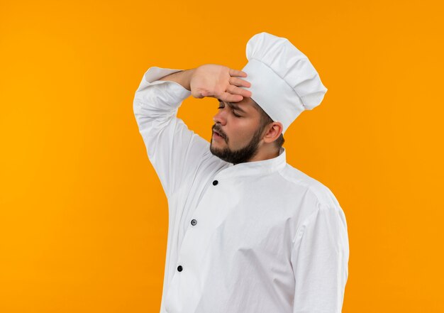 오렌지 공간에 고립 된 닫힌 눈으로 이마에 손을 넣어 요리사 유니폼에 젊은 남성 요리사를 강조