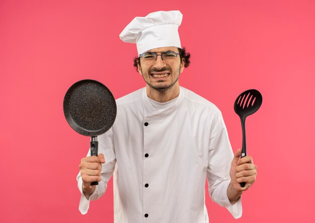 요리사 유니폼과 핑크에 프라이팬과 주걱을 들고 안경을 착용하는 젊은 남성 요리사를 강조