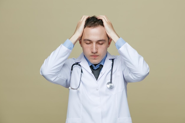 Free photo stressed young male doctor wearing medical robe and stethoscope around neck keeping hands on head with closed eyes isolated on olive green background
