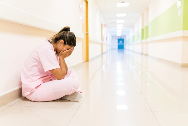 Стрессовая молодая медсестра закрывает лицо, сидя на полу в коридоре больницы