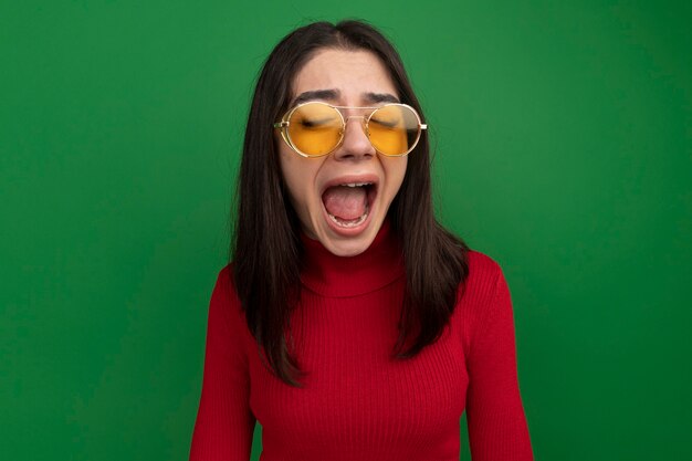 stressed young pretty caucasian girl wearing sunglasses screaming with closed eyes isolated on green wall with copy space