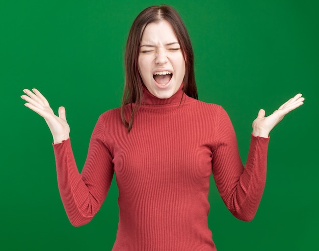 Stressed young pretty woman showing empty hands screaming with closed eyes isolated on green wall