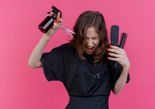 Stressed young slavic female barber wearing uniform holding spray bottle combs and scissors screaming with closed eyes isolated on pink background