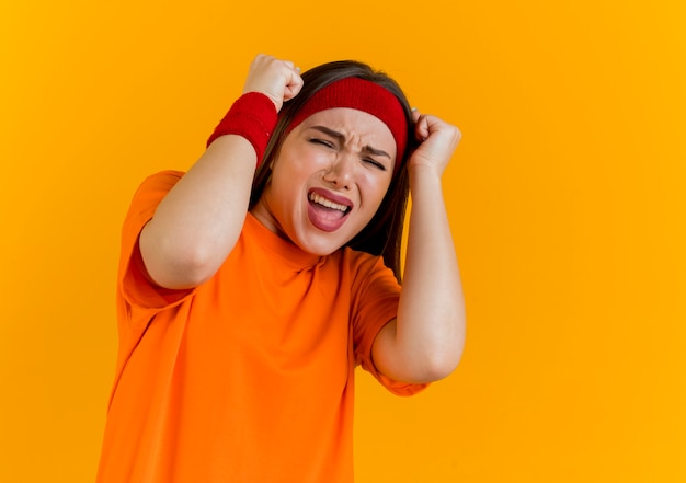 Stressed young sporty woman wearing headband and wristbands touching head with fists screaming with closed eyes 