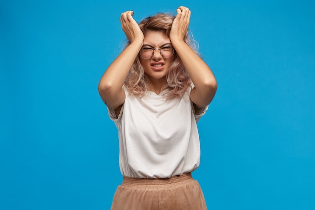 Foto gratuita ha sottolineato la giovane donna in occhiali alla moda che stringe la testa, non sopporta un intollerabile mal di testa a causa della stressante giornata di lavoro. femmina frustrata infastidita che fa smorfie dal dolore