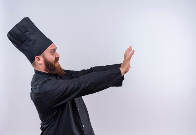 A stressful bearded chef man in black uniform showing stop gesture while looking side on a white wall