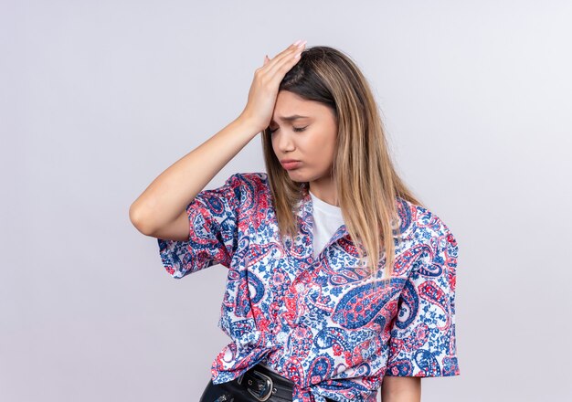 A stressful beautiful young woman wearing paisley printed shirt keeping hand on head on a white wall