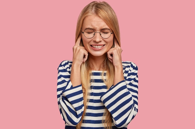 Free photo stressful blonde woman plugs ears, makes wry face, ignores unpleasant sound