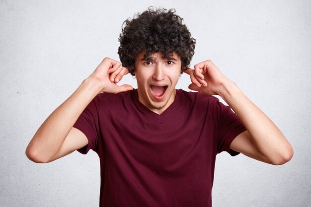 Stressful curly male plugs ears, being in panic, avoids loud irritating sound