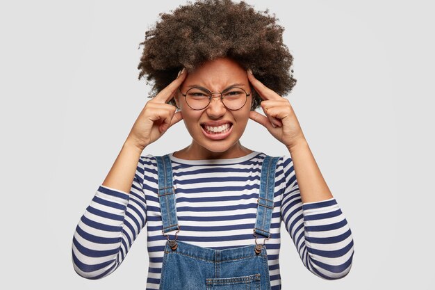 Stressful dark skinned female suffers from terrible headache, keeps fore fingers on temples, clenches teeth with pain, dressed in striped sweater and denim overalls, poses against white wall