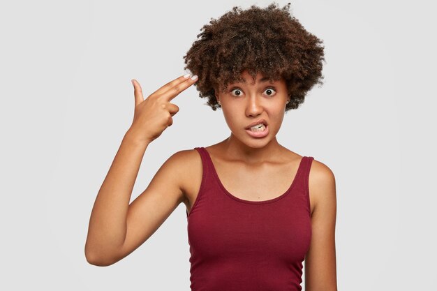 Free photo stressful dark skinned woman with afro haircut, makes suicide gesture in temple, clenches teeth