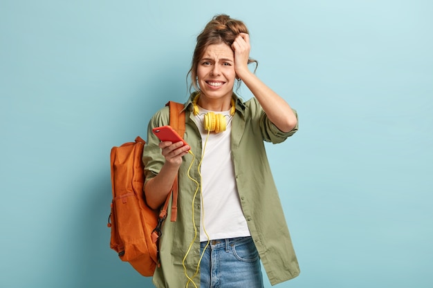 Stressful dissatisfied hipster girl had headache after listening loud music inheadphones for long time, holds modern cell phone