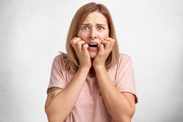 Free photo stressful emotional discontent female model with displeased expression, bites finger nails, looks anxiously, feels worried about something, isolated over white