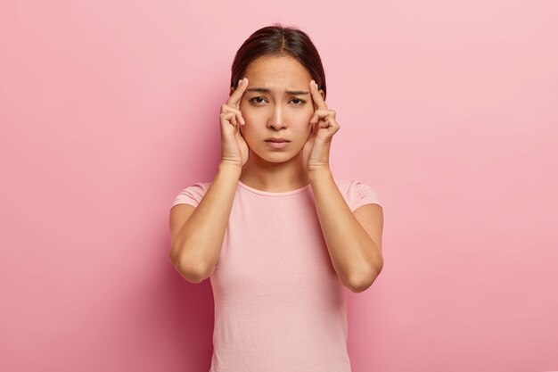 Stressful korean woman keeps fingers on temples, suffers from headache, has painful cramps, looks unhappily , has dark hair, wears casual outfit, isolated on pink wall, feels tired