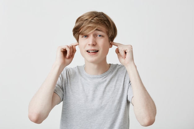 Stressful man with trendy hairdo, dressed casually, plugs ears, avoids loud sounds, doesn`t want to hear noise, isolated against gray background. Fair-haired guy ignores something unpleasant