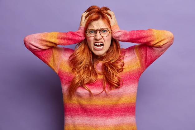 ストレスの多い赤毛の女性は必死に見え、パニックで手を頭に置き、カジュアルなジャンパーの叫び声に身を包んだ不快な音に悩まされ、口を大きく開いたままにします。否定的な感情