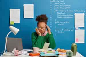 Free photo stressful student feels unwell, has dizziness and headache, unable to work, writes down list to do in notepad, poses against blue background with written information.