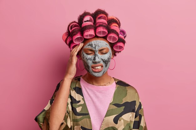 Stressful young woman wears beauty mask and hair curlers, suffers migraine, clenches teeth and closes eyes, tired after loud noisy party, poses at home with displeased expression, models indoor