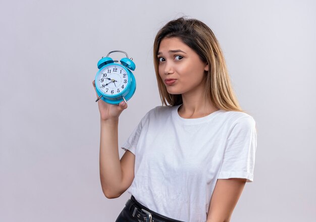 A stressful young woman in white t-shirt showing blue alarm clock 