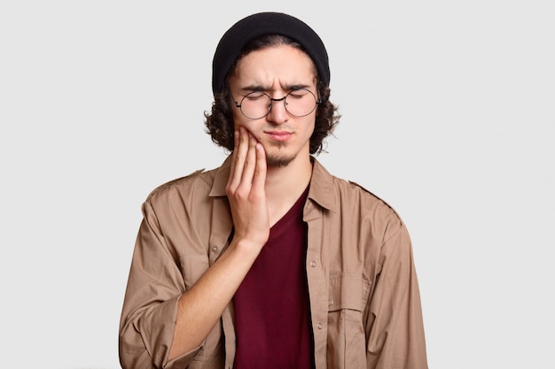 stressful youngster with small beard keeps hand on cheek, suffers from toothache, keeps eyes shut, dressed in stylish clothes, big round spectacles, models on white wall.