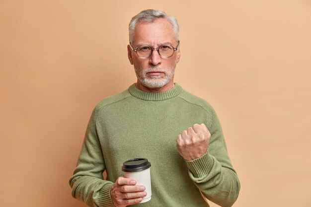 厳格な怒っているひげを生やした年配の男性が真剣に正面を見て、茶色の壁に対してカジュアルなジャンパーポーズを着ている使い捨てのコーヒーを持っていることを警告しようとします