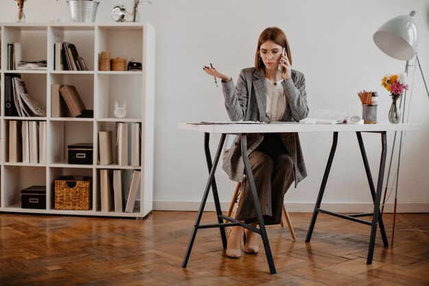 Strict business woman negotiates with clients by phone, sitting in her bright office.