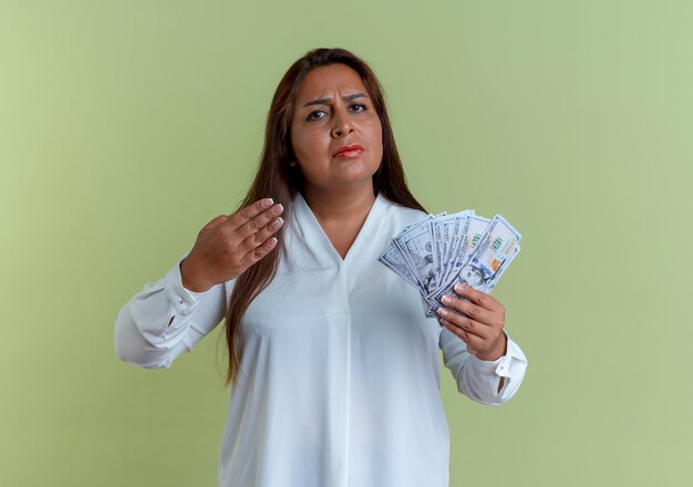 Strict casual caucasian middle-aged woman holding and points with hand at money isolated on olive green wall