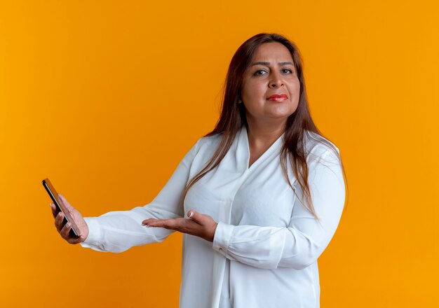 Strict casual caucasian middle-aged woman holding and points with hand to phone isolated on yellow wall