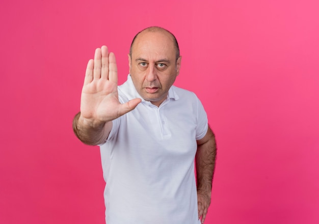 Strict casual mature businessman keeping hand on waist and doing stop gesture at camera isolated on pink background with copy space